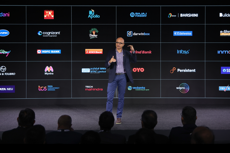 a man wearing a blue jacket delivering a keynote with the screen behind him showing logos of various companies