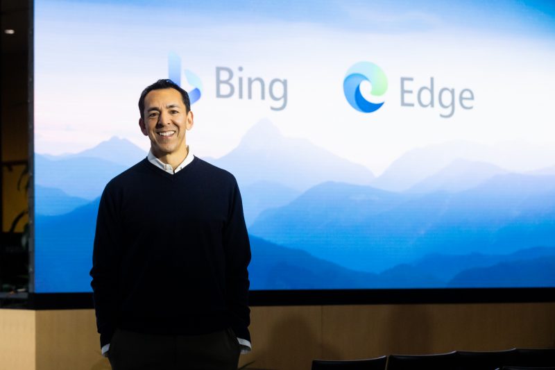 Man smiles as he stands in front of a large screen with Bing and Edge logos.