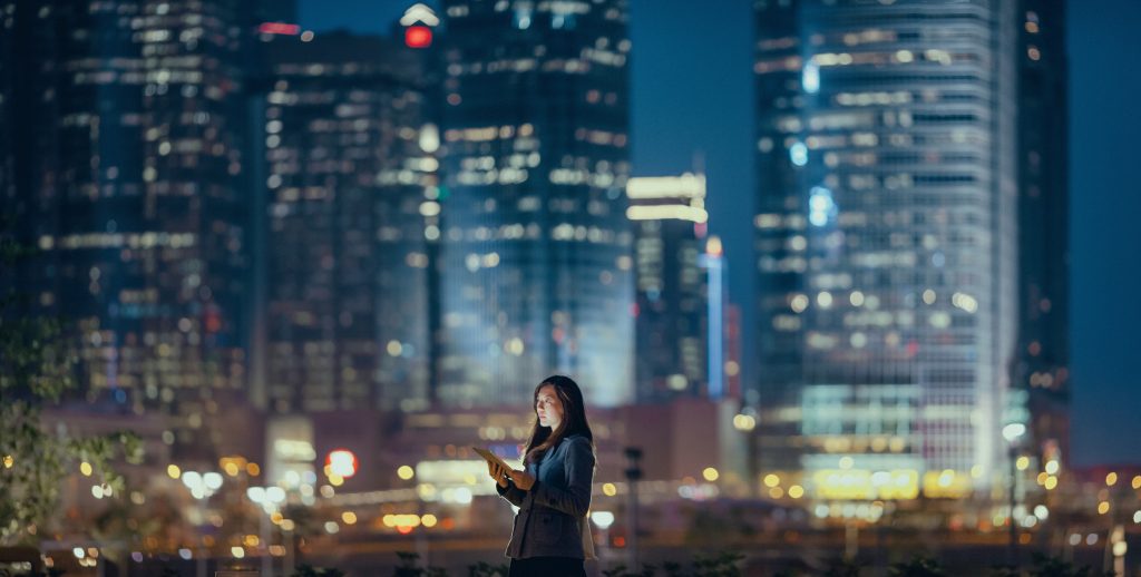 Young businesswoman using digital tablet in financial district, against illuminated corporate skyscrapers at night