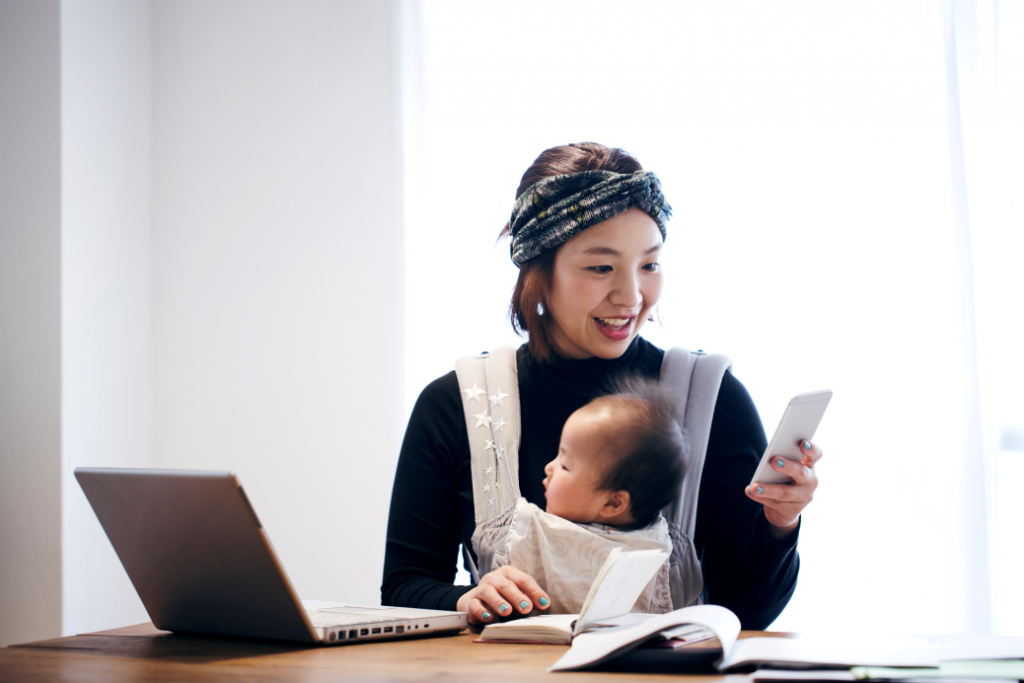 Japanese woman in casual clothes carrying her baby in baby carrier and using a smartphone on the desk.