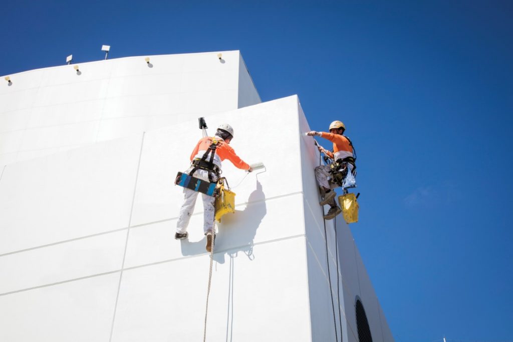 Two workers in safety gear harnessed in high ropes painting a tall building