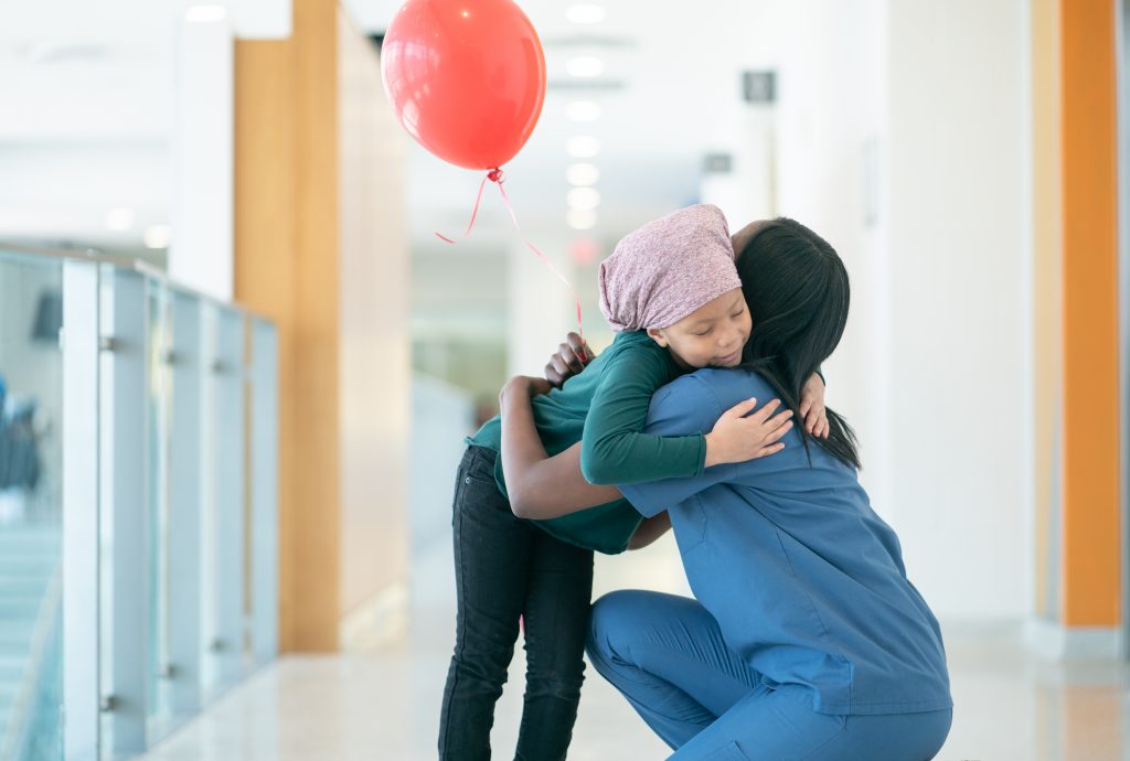 A young female cancer patient wearing a headscarf gives her nurse a big hug as they are both at the hallways at the hospital. She is also holding a red balloon that was gifted to her from the hospital staff.