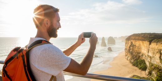 Cheerful young man takes a picture using a mobile phone of the Twelve Apostles sea rocks on the Great Ocean Road in Victoria's state of Australia.