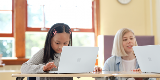 Elementary school age child sitting in a classrom with an open Surface Laptop SE in Glacier in front of her. A teacher is also shown.