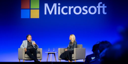 A Caucasian male and female sitting across from one another in seated lounges, on stage, with a Microsoft logo behind them