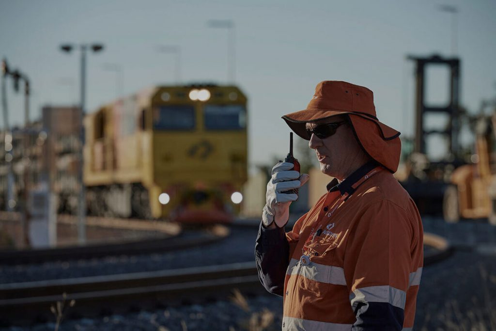 Adult male in safety uniform talking in a walkie talkie, with a rail train in the background
