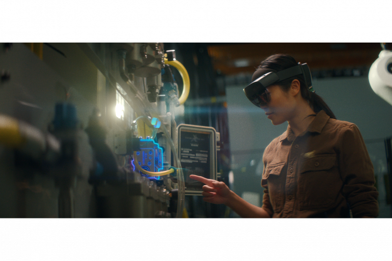 Woman in an industrial setting, wearing a HoloLens 2, pointing at a component on a hydraulic panel.
