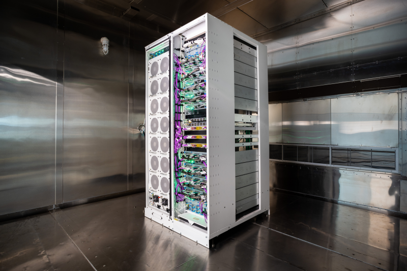 A rectangular piece of equipment roughly the size of a large refrigerator with a column of datacenter servers next to a column of fans sits inside a shiny metal room where temperature can be controlled.