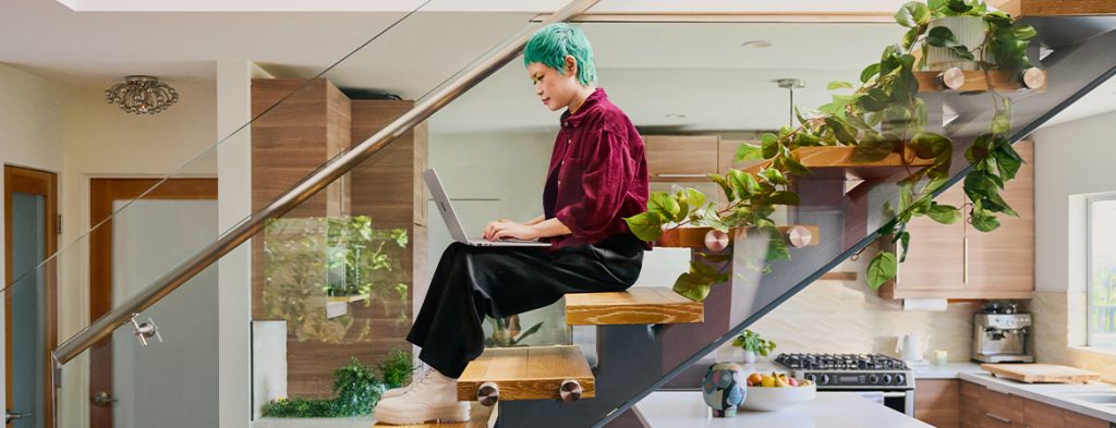 Una persona está sentada en unas escaleras con plantas en macetas junto a la cocina, trabajando con un portátil.