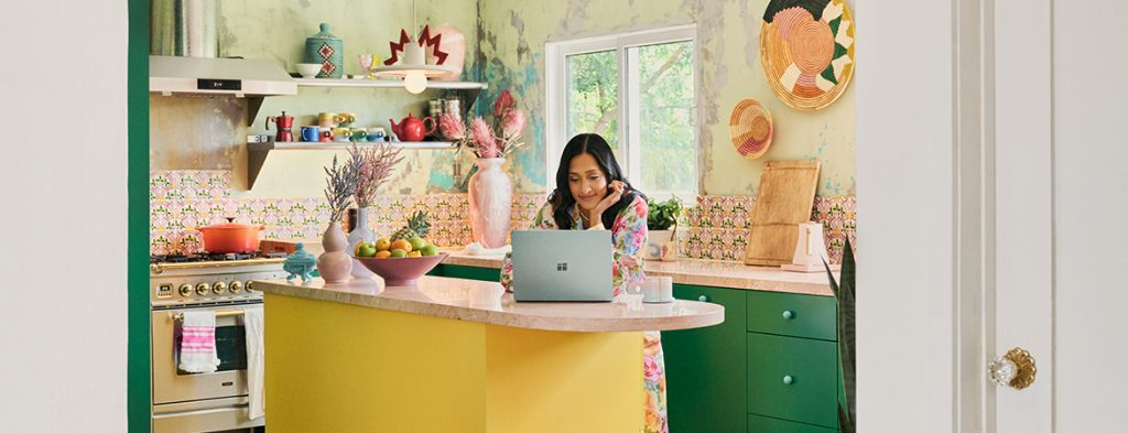 Uma mulher está numa ilha de cozinha a trabalhar com um computador portátil.
