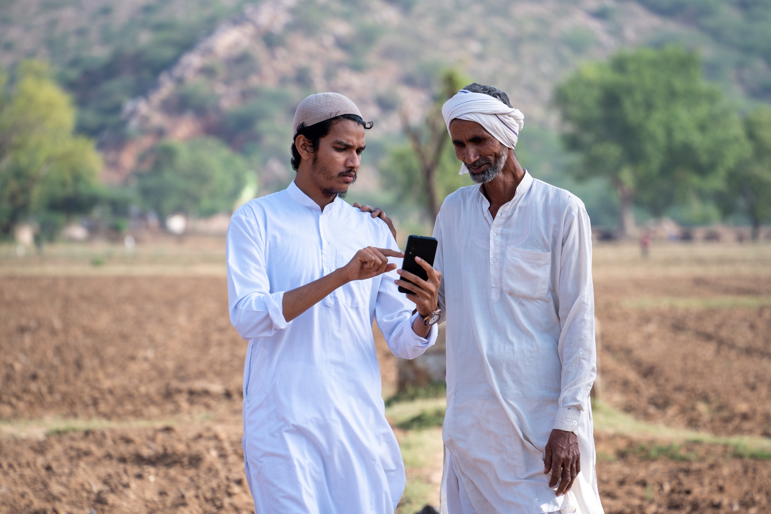 Two male villagers standing in an arid Indian field looking at a smartphone