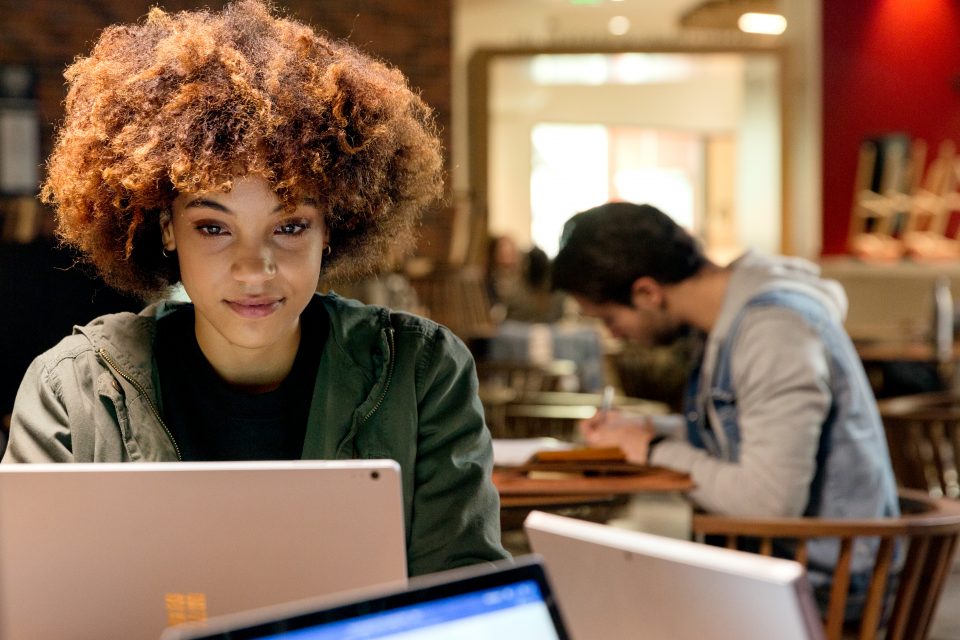 Close up of female college student sitting at table in study café, using Surface Book (screen not shown). Two other laptops are also in use at table she shares.