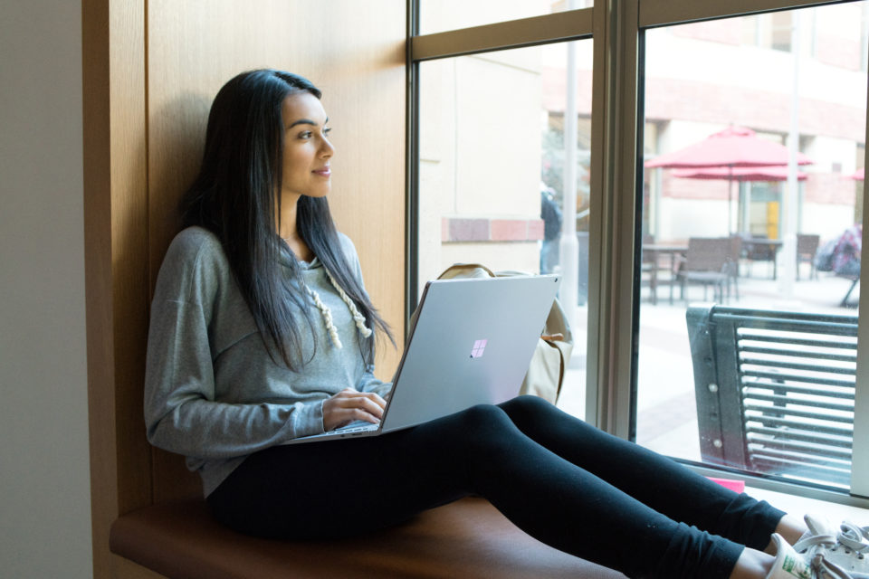 Female college student sitting in windowsill and looking out window at campus. An open Surface Book sits on her lap