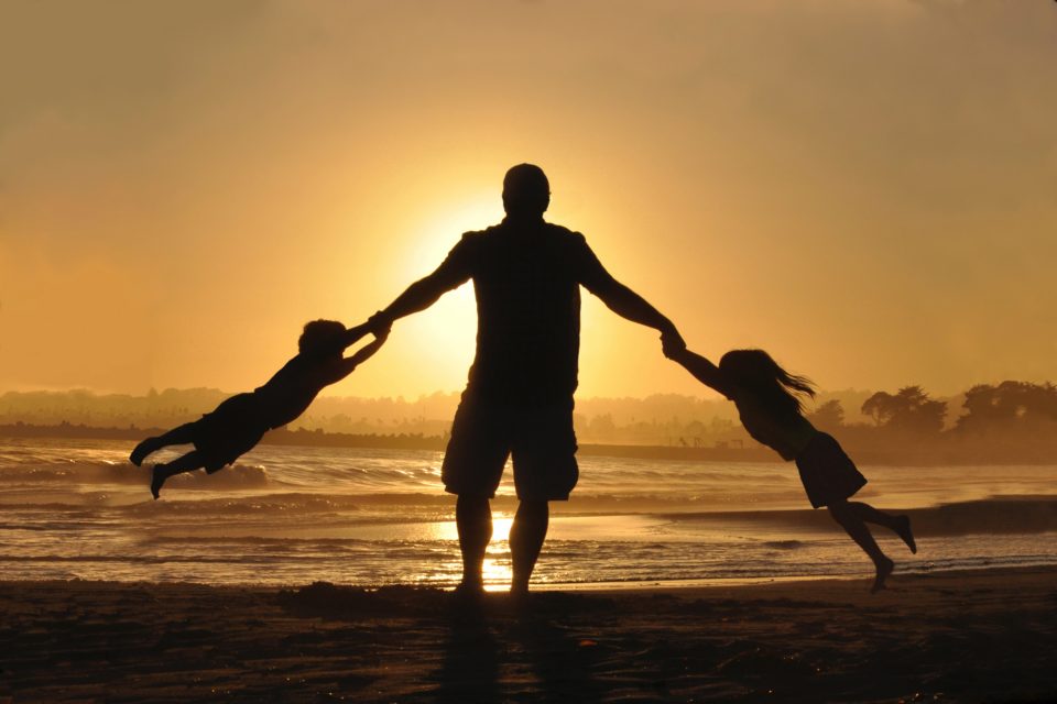 Man on beach at dusk, with kids swinging on his arms