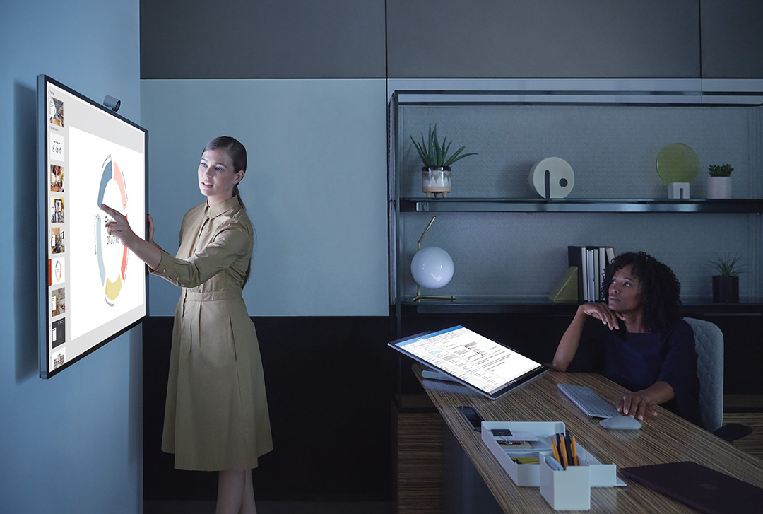 A woman uses a Surface Hub 2S that is mounted to a wall in an office