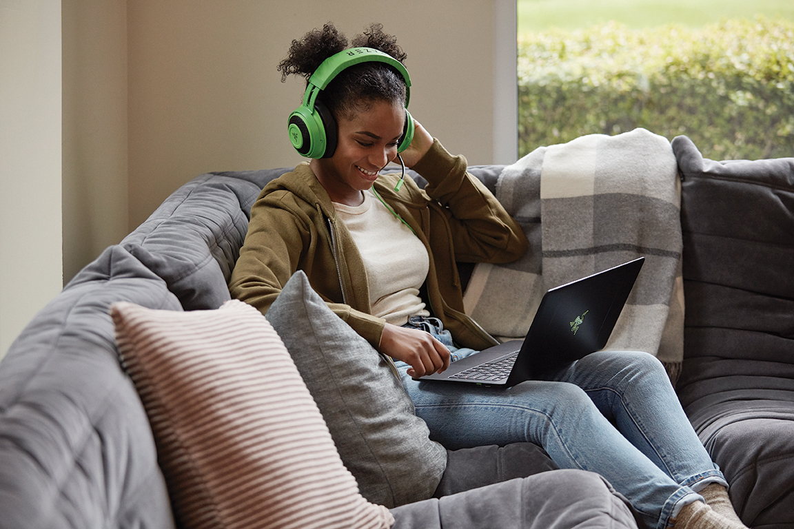 A woman wearing gaming headphones sits on a sofa at home with a laptop open on her lap