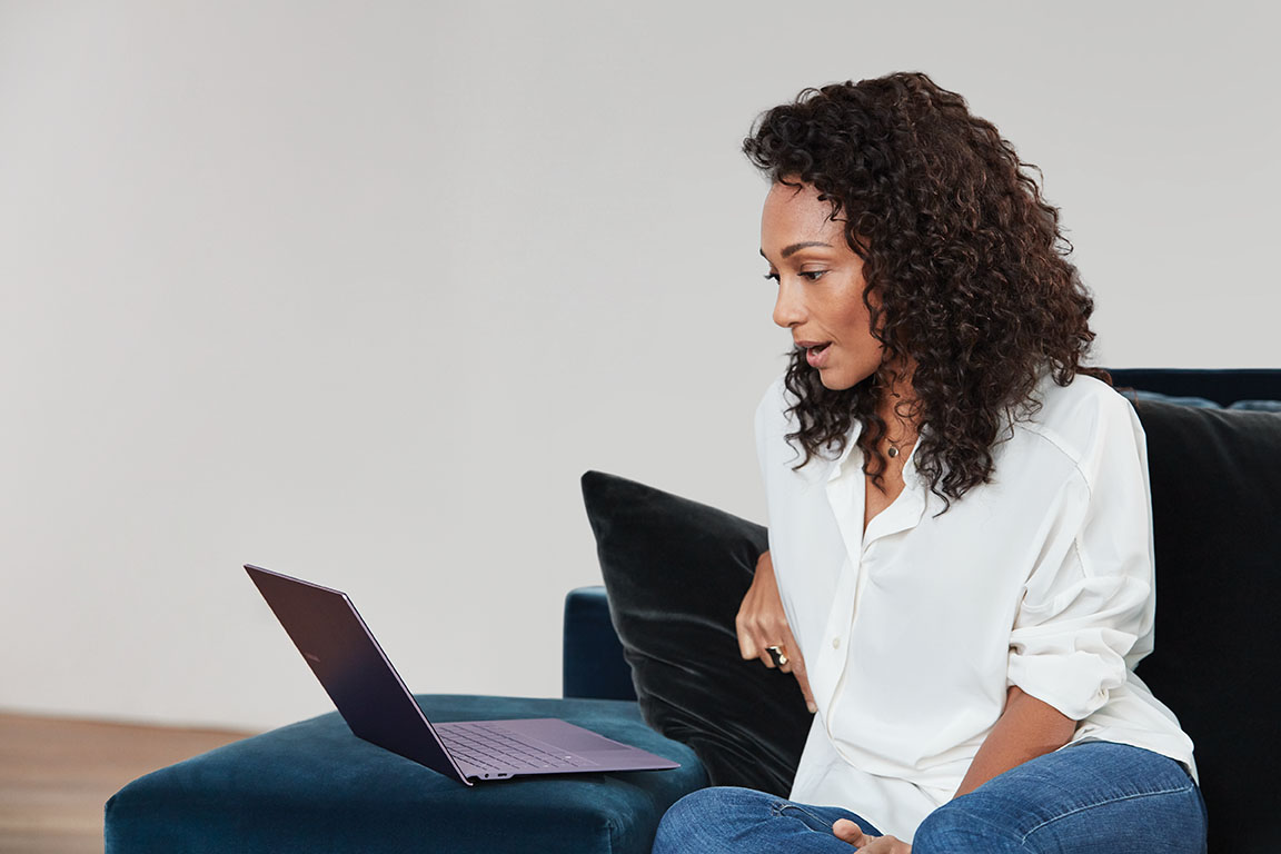 A woman sitting on a sofa looks at a Surface laptop that's open in front of her