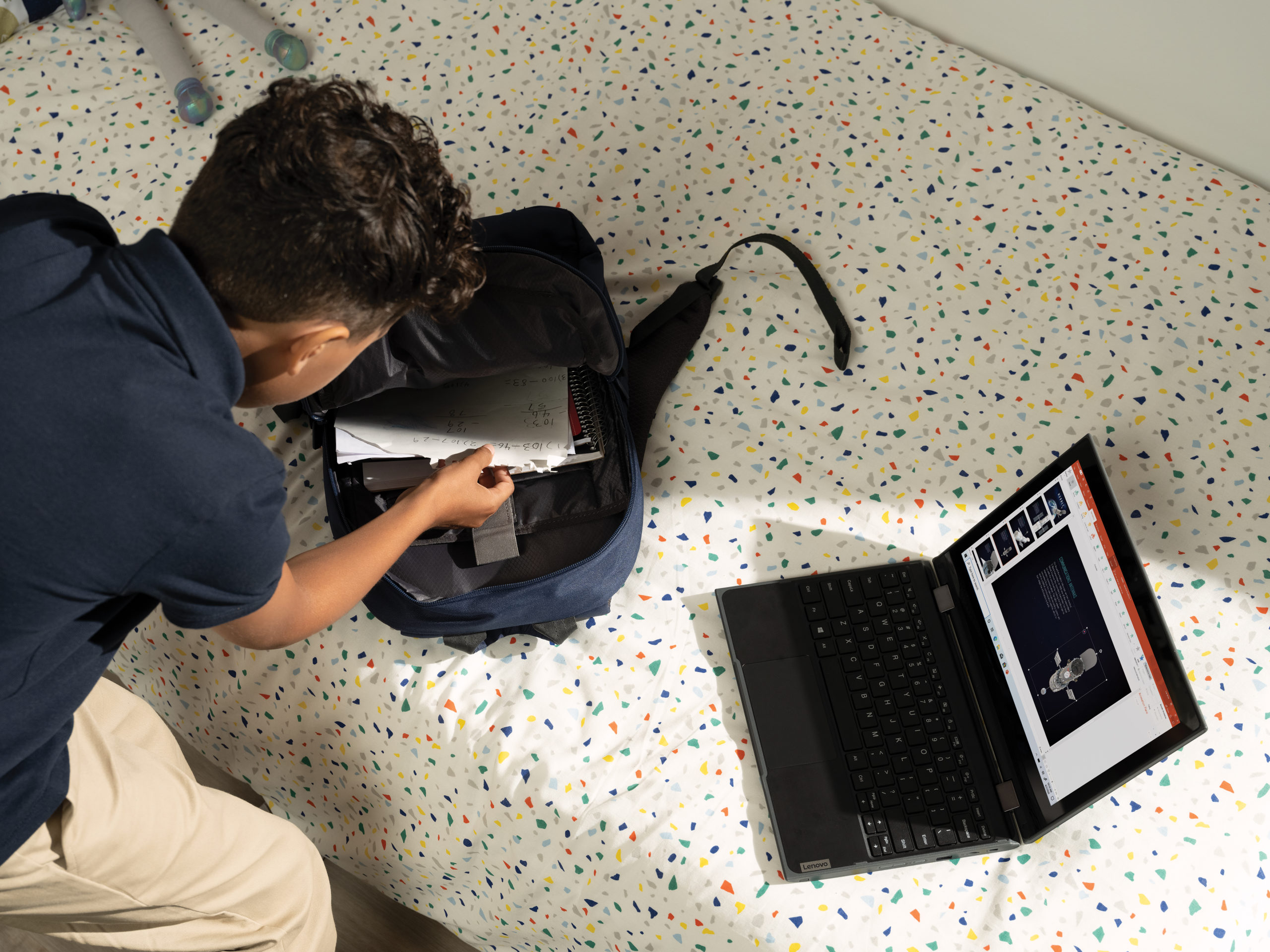 A middle school boy loading up his backpack in his bedroom with a Lenovo 300e 2nd Generation 2-in-1 opened in laptop mode displaying a PowerPoint file. Remote Learning collection.
