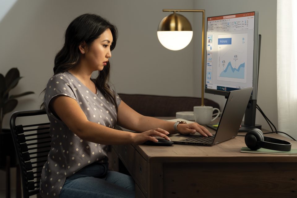 A woman working at her home office on a Lenovo ThinkPad Yoga connected to an external monitor displaying a PowerPoint file