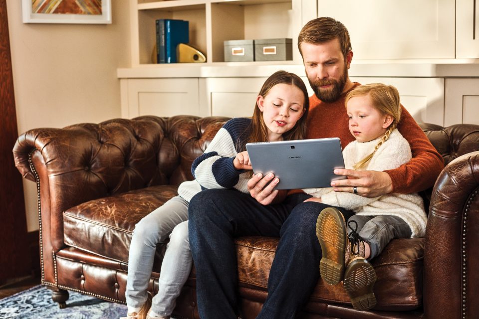 Man sits on sofa at home with two young daughters, looking at tablet device