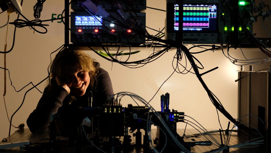 A woman is looking out underneath some computers with a lot of wires and lights. She is smiling with her head in her hand.