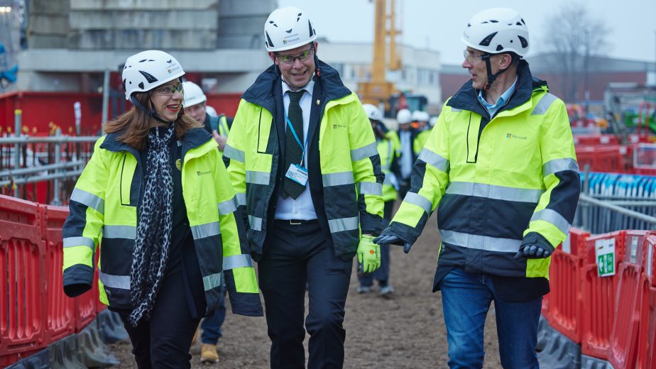 Clare Barclay, CEO of Microsoft UK, John Madden, and Chancellor Jeremy Hunt visit the new Microsoft data centre that is being built in west London. The photo shows the three of them walking outside the building site while wearing hard hats and high-vis jackets.