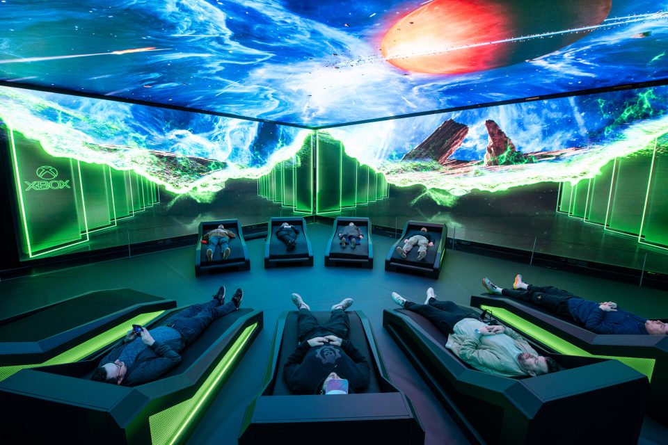 A photo of the Xbox Power Your Dreams immersive experience in London. It shows people lying on individual bed pods as they are surrounded by HD images of gaming scenescapes on the ceiling and walls