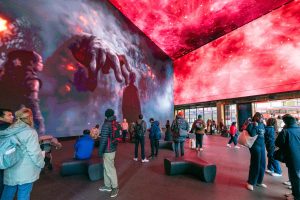 A photo of people in the main hall of the Outernet, which shows HD gaming scenescape imagery across the walls and ceiling. In this picture, the imagery is of a large hand pointing toward a person, as they are surrounded by red and grey galaxies.