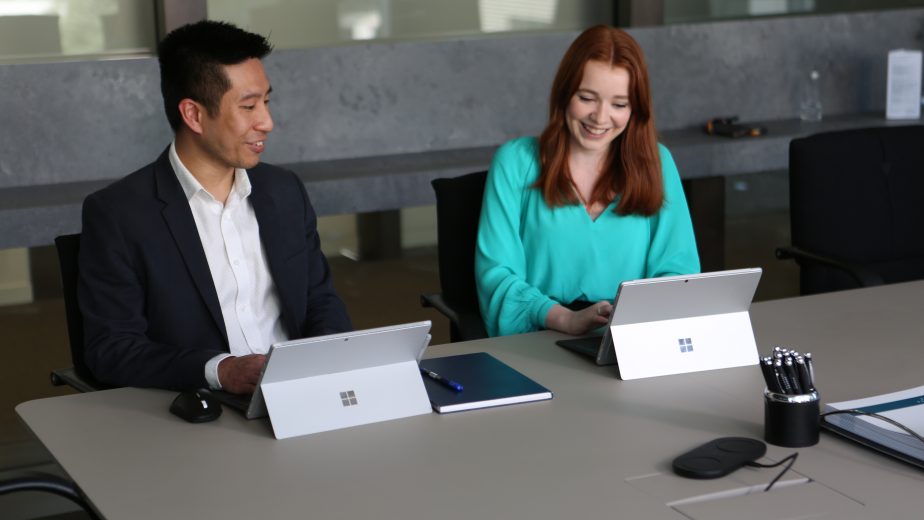 A man and a woman in an office sit at a desk with a Surface device each. They are smiling as they do work on their Surface devices.