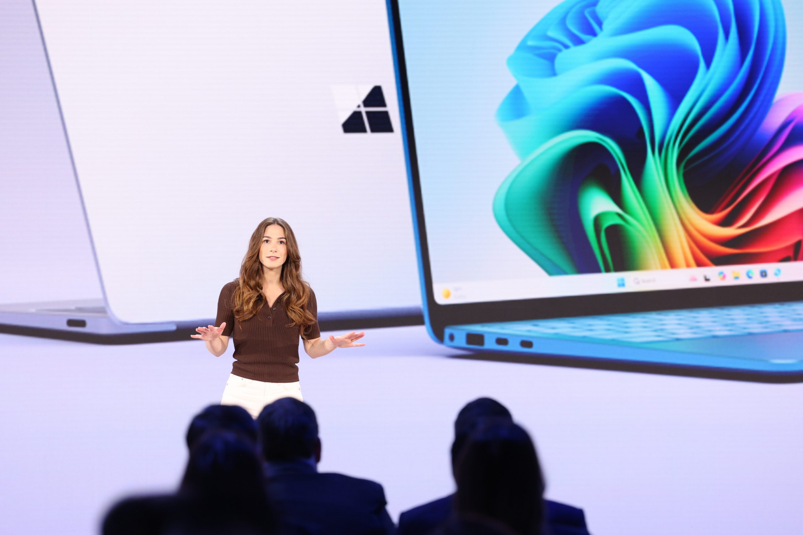 A woman standing on stage in front of an audience, with an image of a laptop on the screen behind her