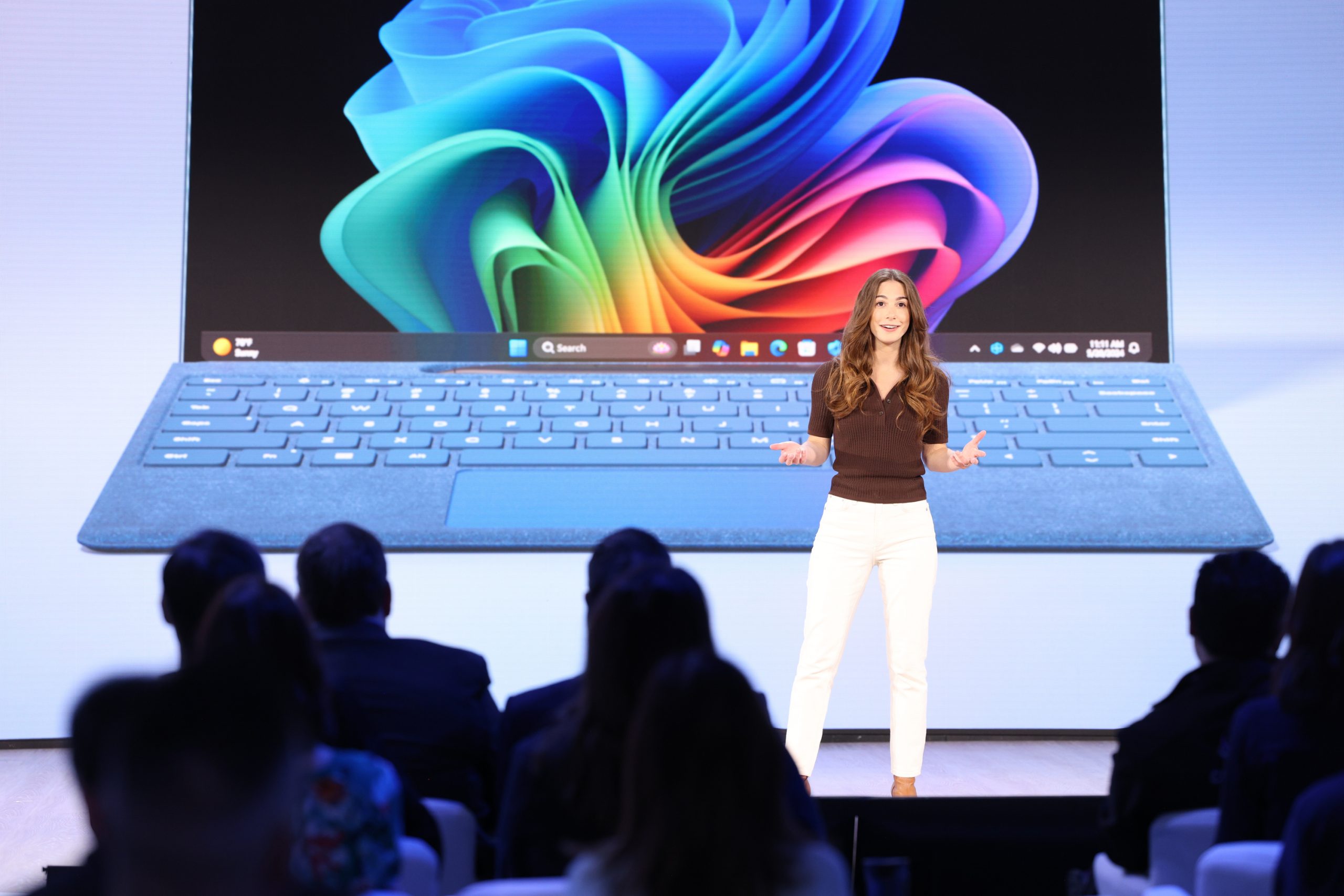 A woman standing on stage in front of an audience, with an image of a laptop on the screen behind her.