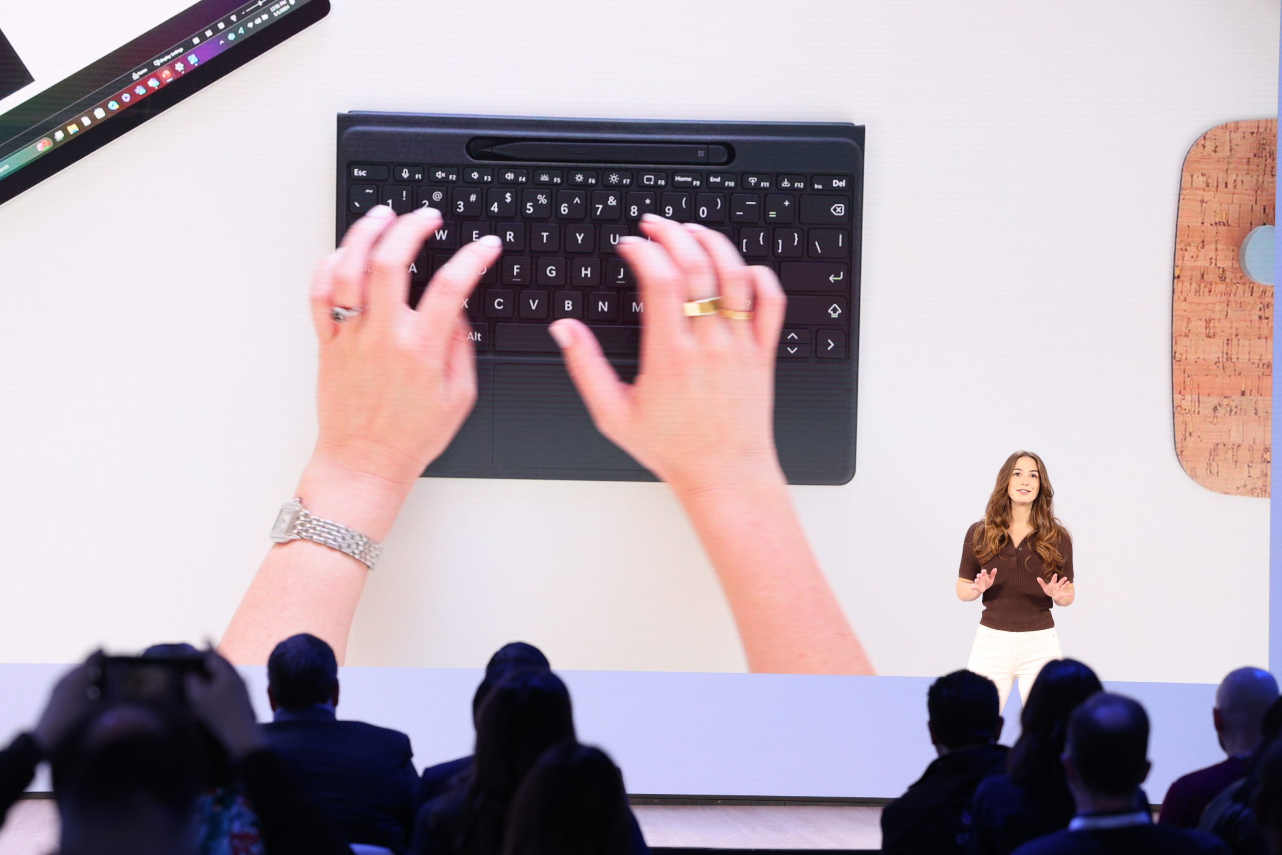 A woman standing on stage in front of an audience, with an image of hands typing on a keyboard on the screen behind her.