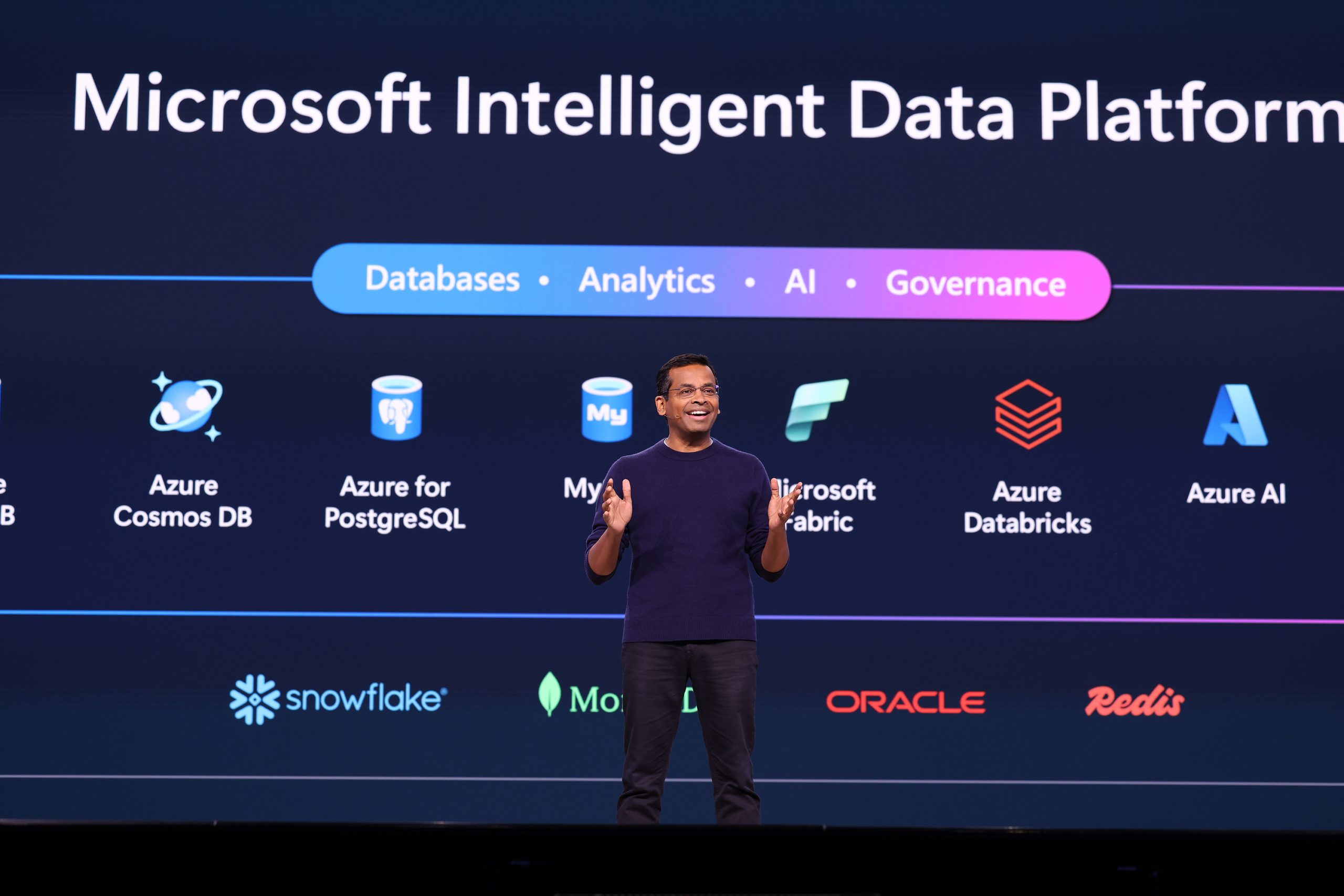A man standing on stage speaking to an audience and gesturing with his hands, with text on the screen behind him about the Microsoft Intelligent Data Platform