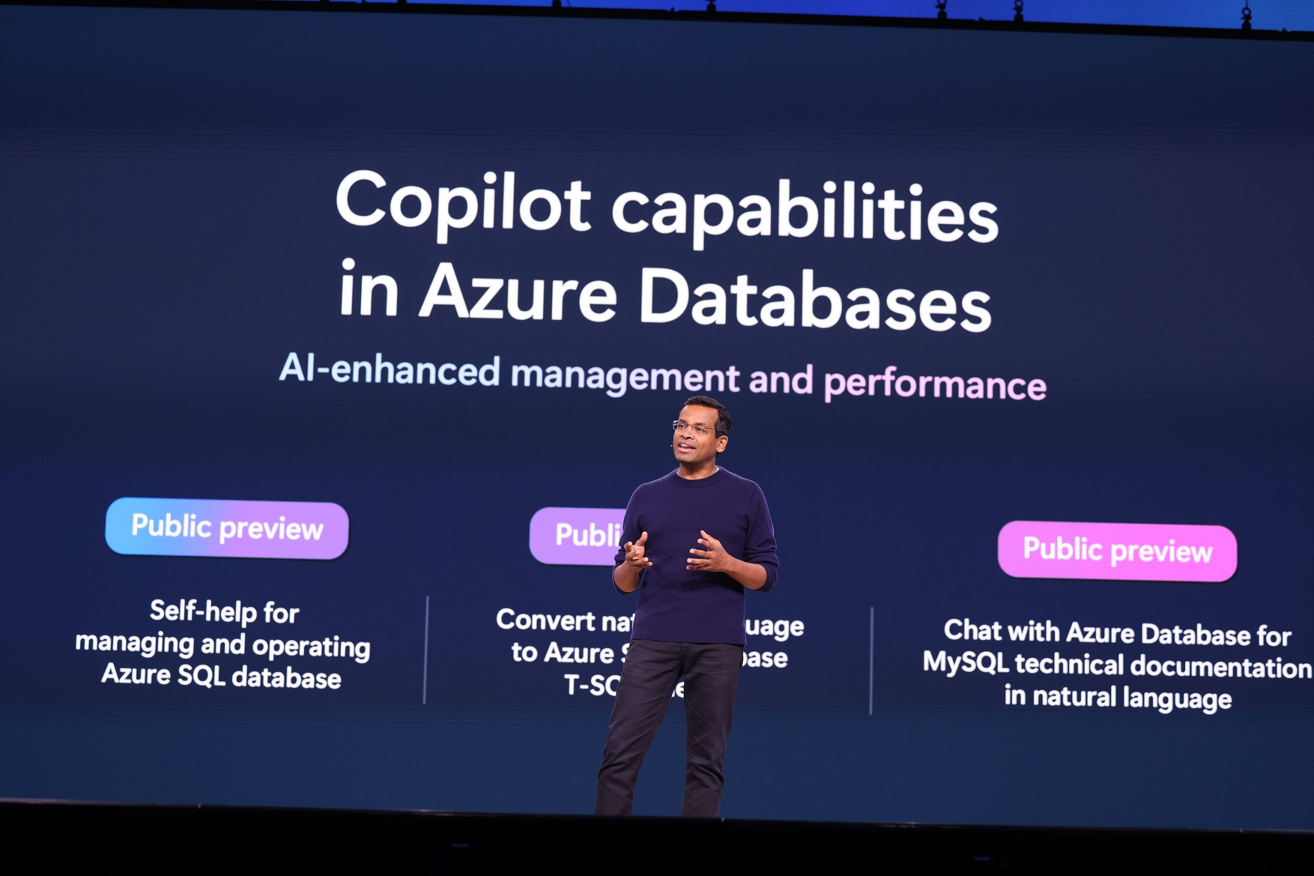 A man standing on stage speaking to an audience and gesturing with his hands, with text on the screen behind him about Copilot capabilities in Azure Databases