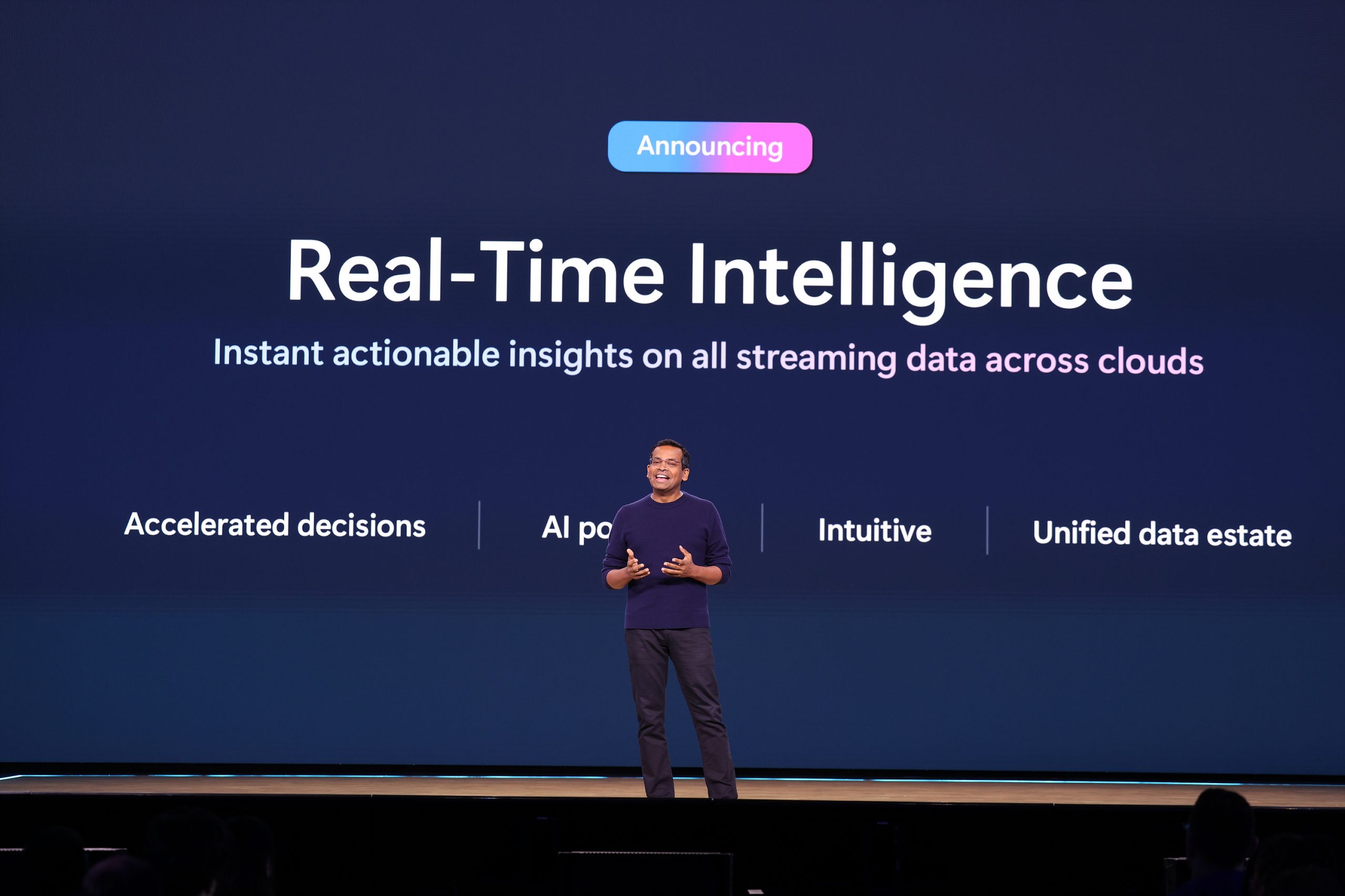 A man standing on stage speaking to an audience and gesturing with his hands, with text on the screen behind him about Real-Time Intelligence