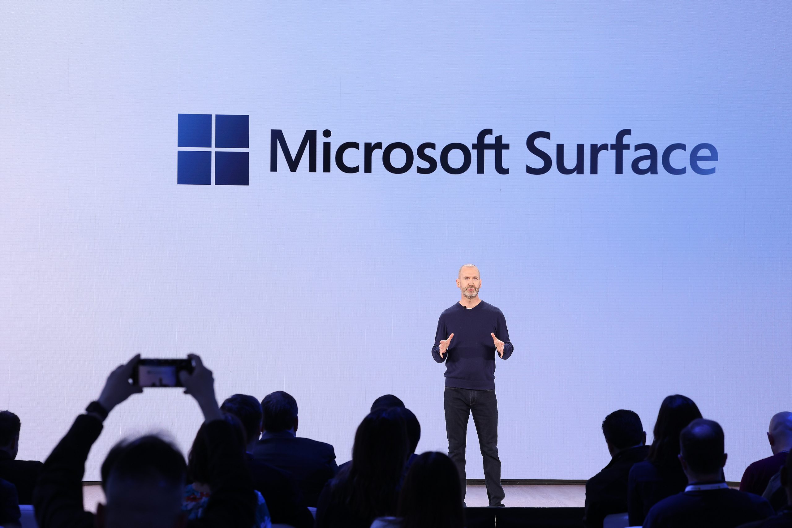 A man standing on stage in front of an audience, with the Microsoft Surface logo on the screen behind him
