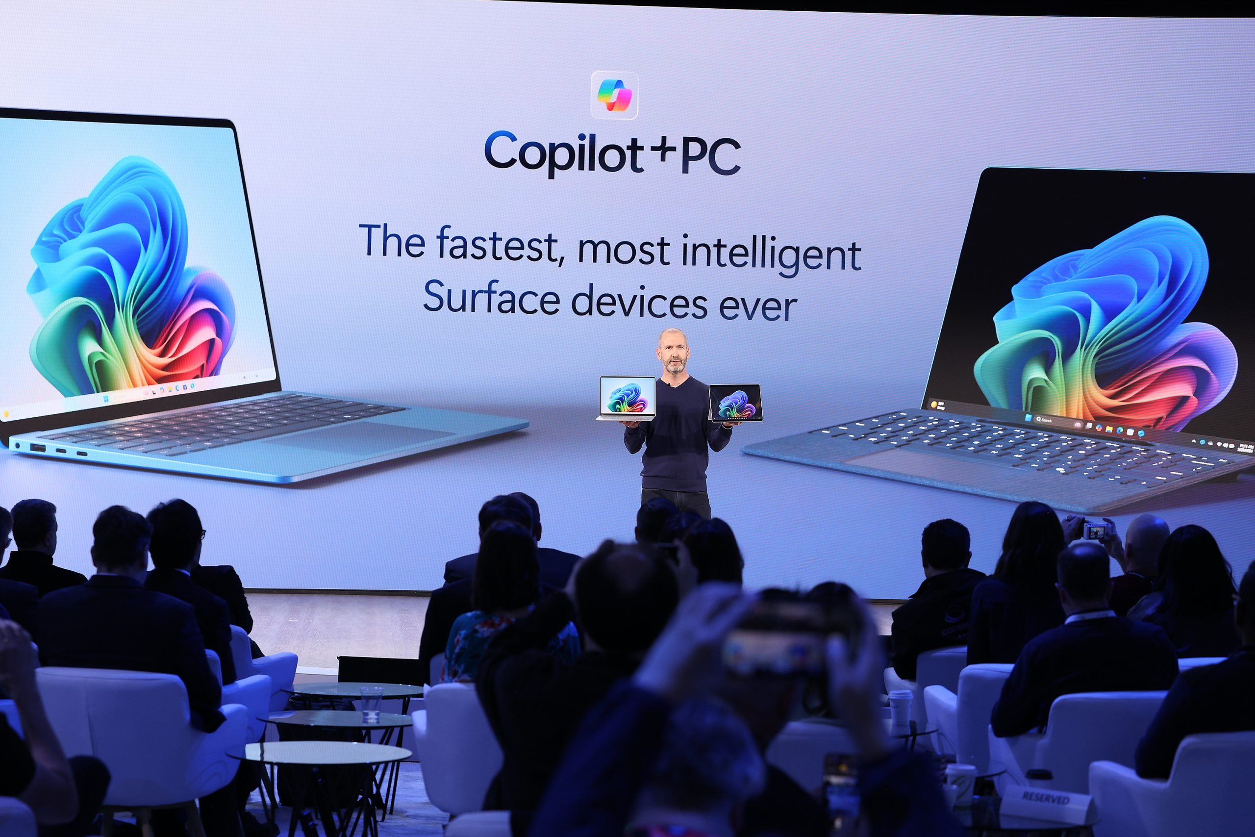 A man standing on stage in front of an audience holding two Copilot+ PCs, in front of a screen that reads, 