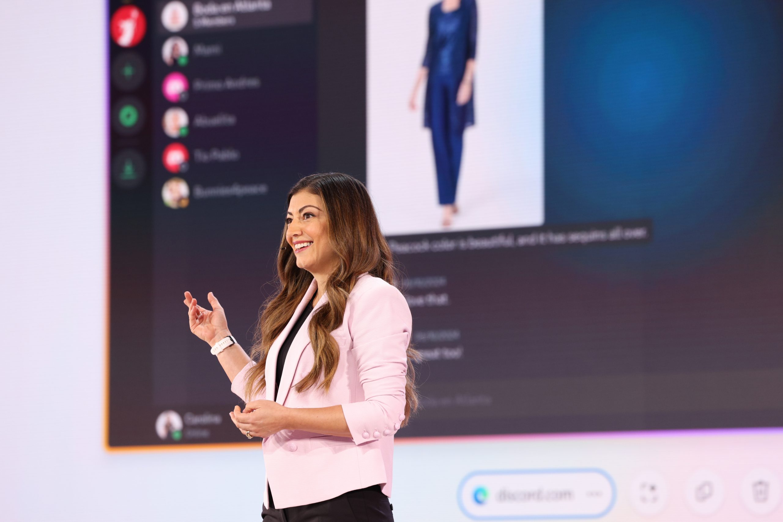 A woman standing on stage in front of an audience, with a device user interface projected on the screen behind her