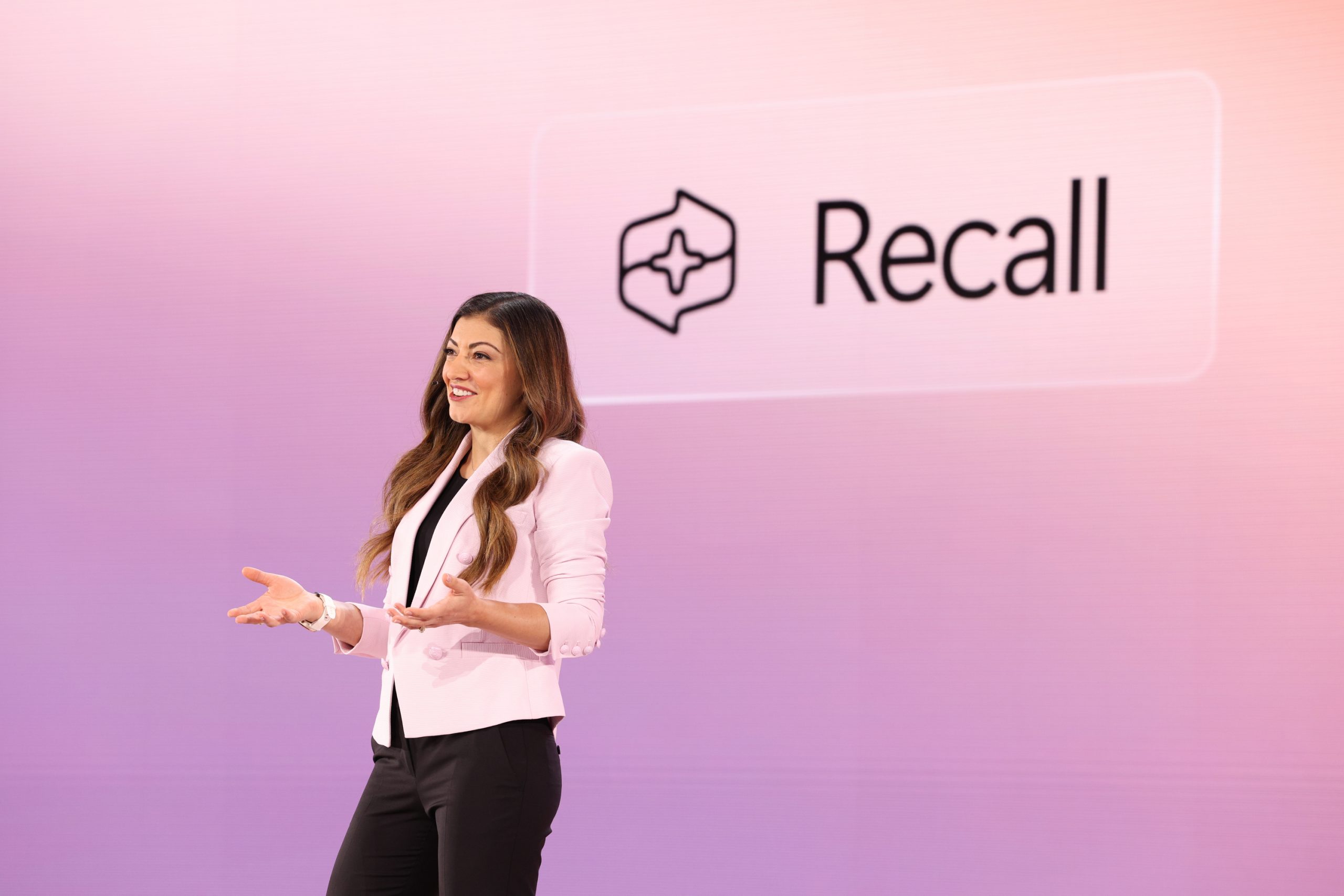 A woman standing on stage in front of an audience, with the Recall word and logo on the screen behind her