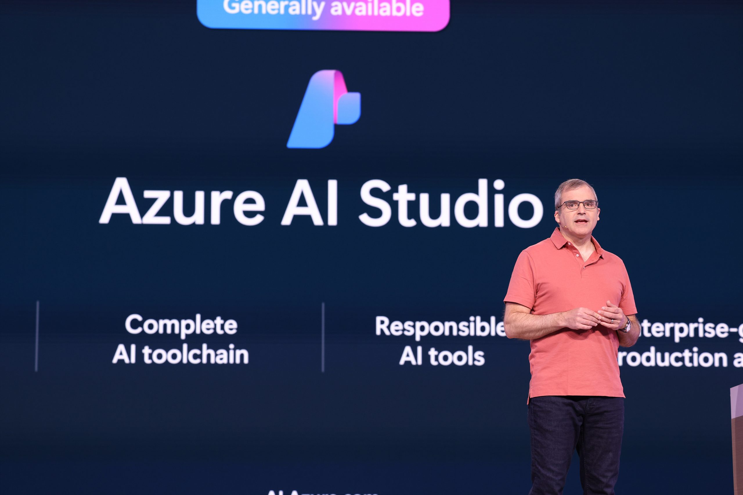 A man standing on stage speaking to an audience with text about Azure AI Studio on a screen behind him