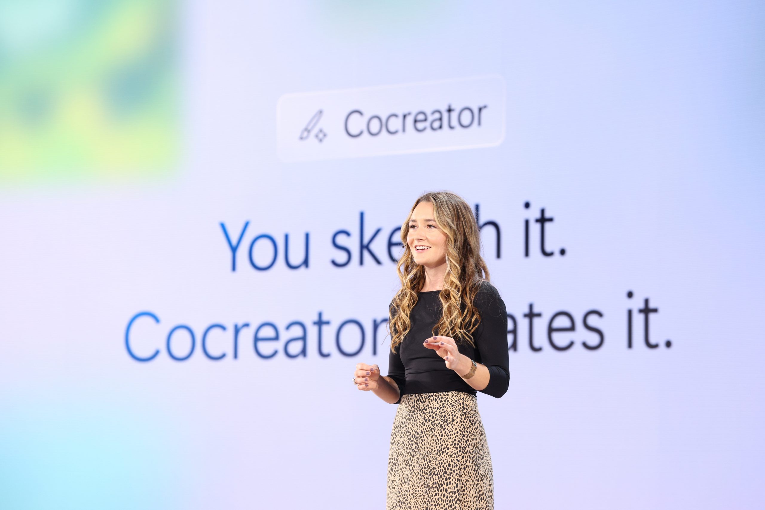 A woman standing on stage in front of an audience with the Cocreator logo and tagline on the screen behind her