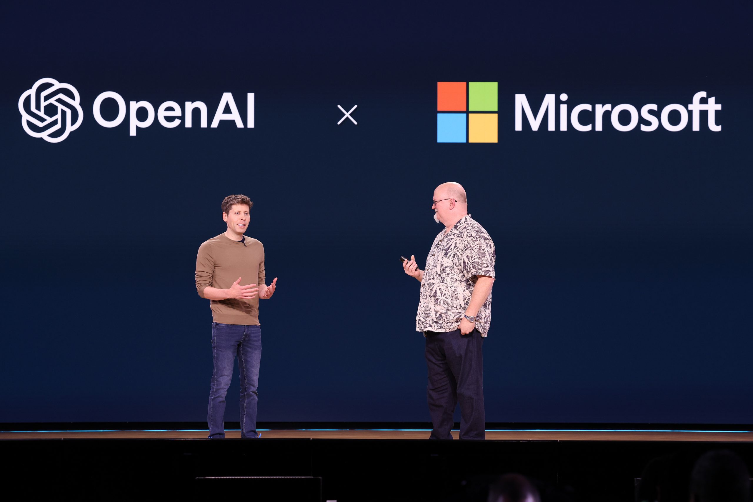 Two men standing on stage with OpenAi and Microsoft logos on a screen behind them