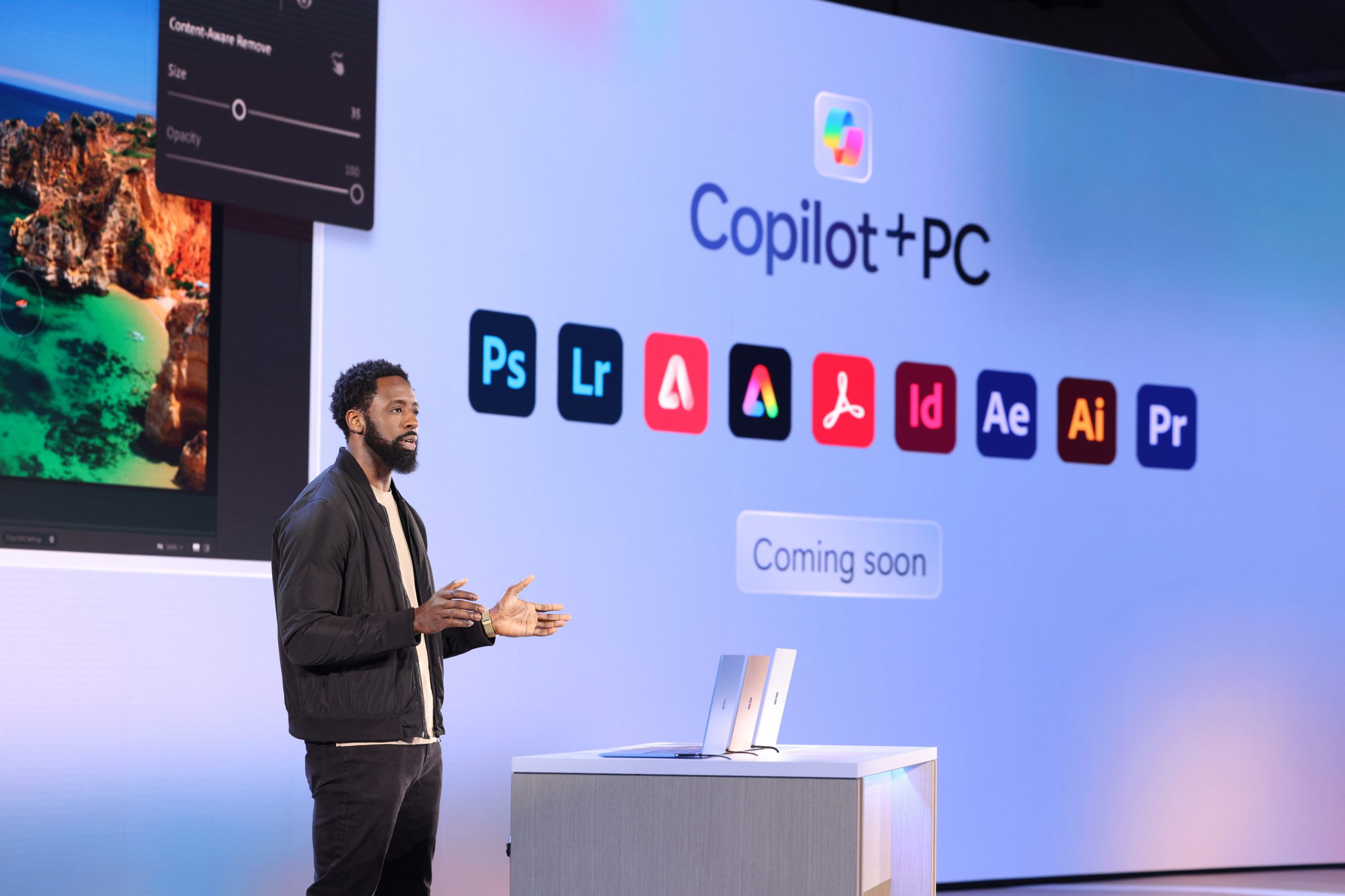 A man standing on stage in front of an audience with a collection of logos on the screen behind him that represent Copilot+ PC