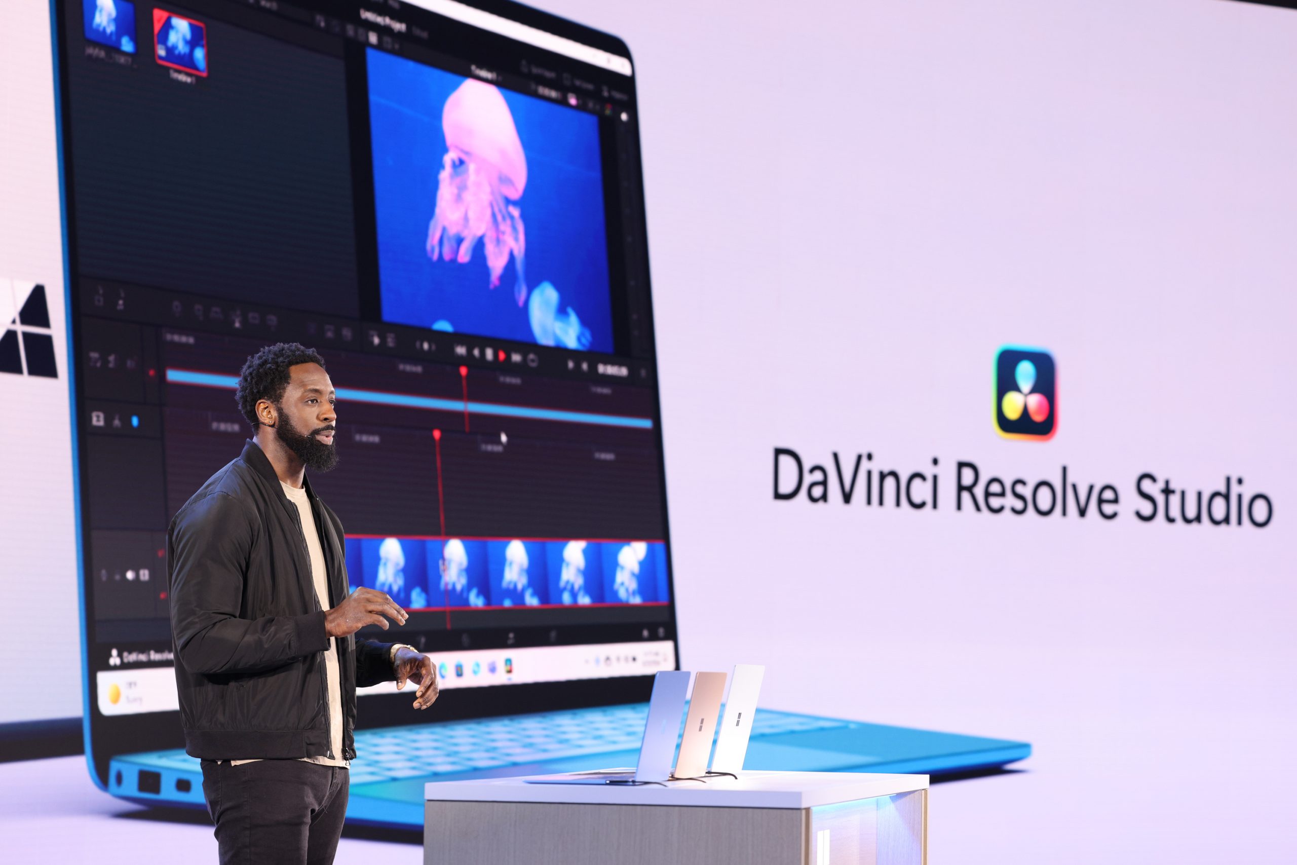 A man standing on stage in front of an audience with a laptop demonstrating DaVinci Resolve Studio projected onto the screen behind him