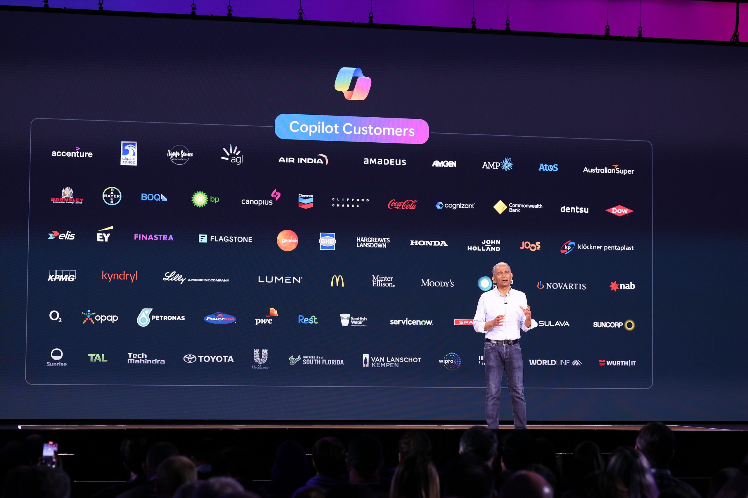 A man standing on stage with a list of Copilot customers on a screen behind him