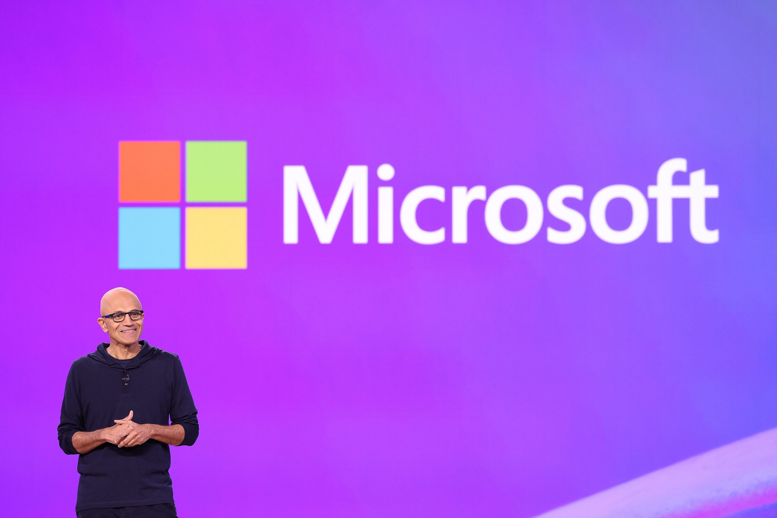 A man standing on stage with a purple background and the Microsoft logo on the screen behind him