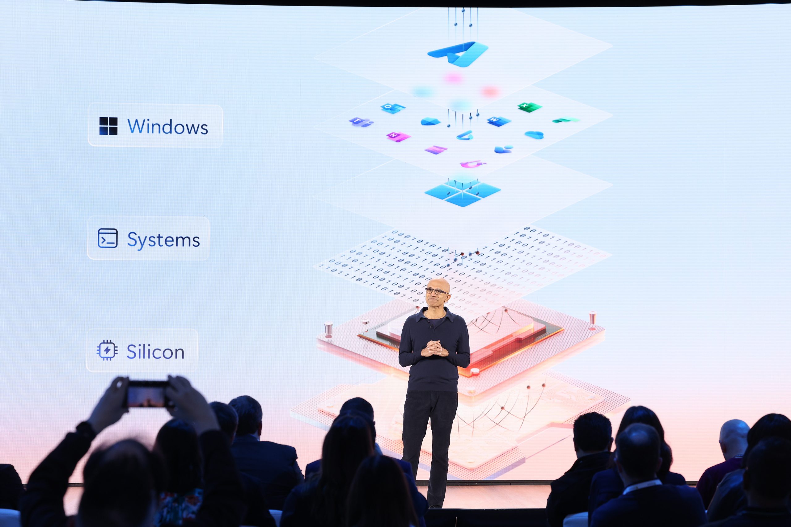 A man stands on a stage in front of an audience next to icons that denote Windows, Systems and Silicon