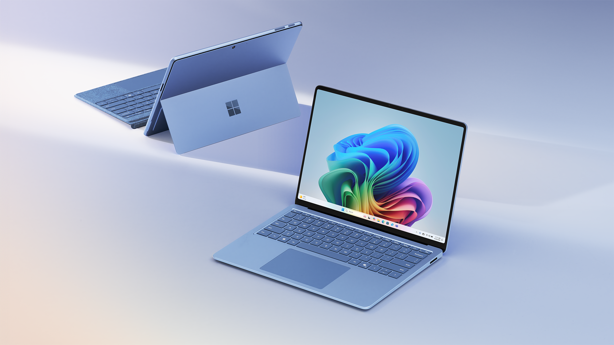 Two Surface devices with an angled back view of a blue Surface Pro including the detached Surface Pro Keyboard and an angled front view of a Surface Laptop opened with the Windows rainbow bloom image on the home screen.