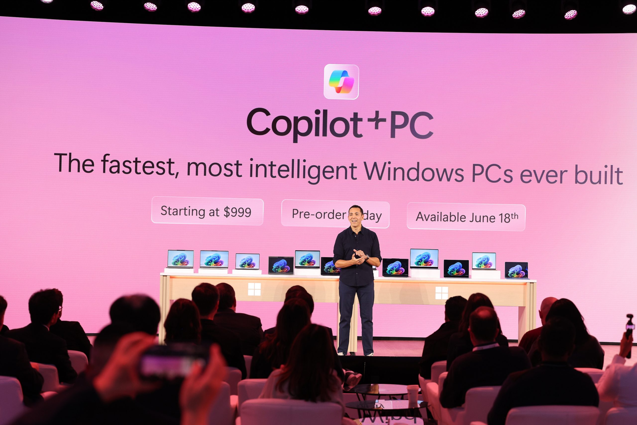 A man standing on stage in front of an audience with a display of Copilot+ PC laptops behind him