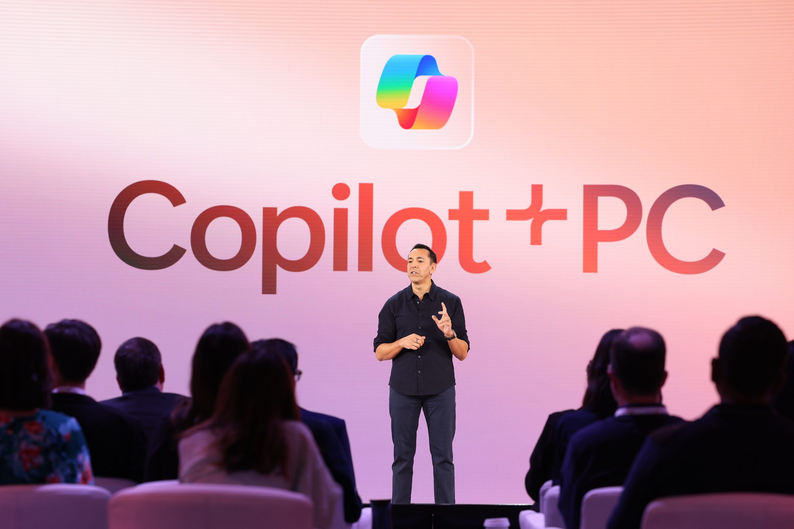 A man standing on stage in front of an audience with the Copilot+ PC logo on a rose-gold screen behind him.